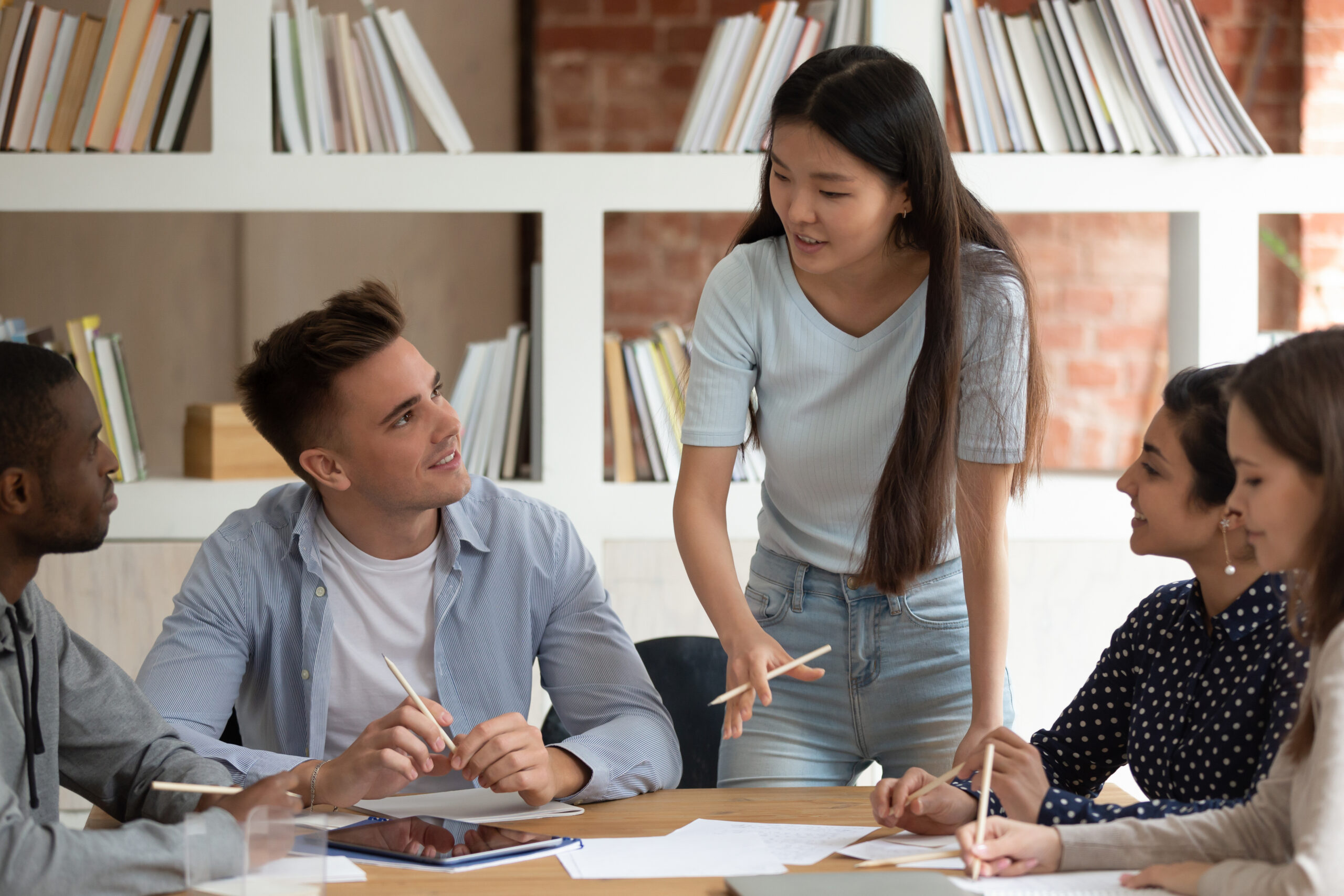 Motivated Asian female team leader stand hold meeting with mates talk discuss project or assignment, focused young ethnic girl speaker or tutor speak with students at group gathering. Teamwork concept